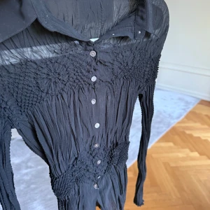 Svart plisserad blus från Marks & Spencer - Säljer en svart långärmad blus från Marks & Spencer med plisserad struktur och smockdetaljer. Klänningen har knappar framtill och klassisk krage. Perfekt för dig som gillar unika detaljer och en figurnära passform.