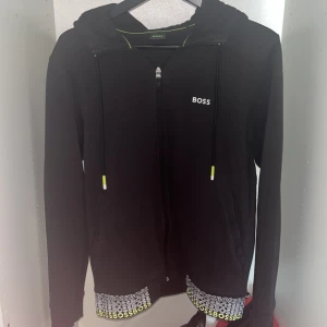 Hugo boss hoodie svart - Sällsynt hoodie med dragkedja från BOSS. Har huva med snören som har neongula och vita detaljer. Nederkanten är dekorerad med broderade BOSS-loggor i vitt och neongult. Perfekt för en sportig och stilren look.