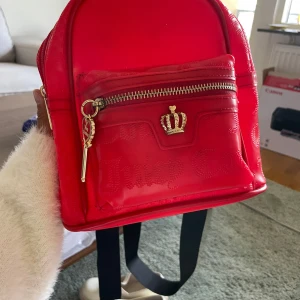 Röd ryggsäck från Juicy Couture - Säljer en snygg röd mini-ryggsäck från Juicy Couture med guldfärgade detaljer och en krona framtill. Väskan har ett ytterfack med dragkedja och justerbara svarta axelband. Materialet är syntet (polyvinylklorid). Perfekt för dig som vill sticka ut!