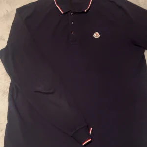 Mörkblå pikétröja från Moncler - Snygg mörkblå långärmad pikétröja från Moncler med klassisk krage och knappar framtill. Tröjan har Moncler-logga på bröstet och diskreta rödvita detaljer på krage och ärmslut.