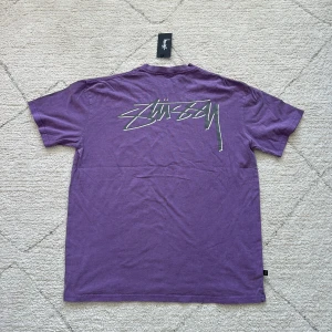 Lila t-shirt från Stüssy - Säljer en lila t-shirt från Stüssy med logga tryckt på bröstet och i stort format på ryggen. T-shirten har rund halsringning och normal passform. Perfekt för dig som gillar streetwear och vill sticka ut med en snygg färg.