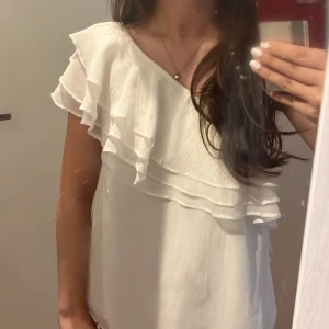 Vit off shoulder topp - Säljer en vit topp med volangdetaljer och asymmetrisk axel. Toppen har en lös passform och är tillverkad i ett lätt, plisserat material. Perfekt för sommardagar när du vill känna dig både stilren och bekväm.