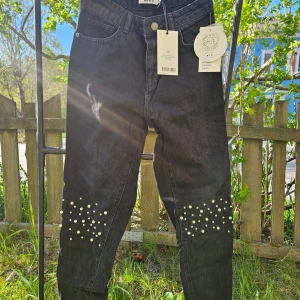 Svarta jeans med pärldetaljer - Snygga svarta jeans med pärlor på knäna som ger en cool och unik look. Byxorna har klassisk femficksdesign och normal passform. Perfekta för dig som vill sticka ut lite extra. 