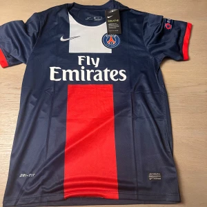 PSG fotbollströja Ibrahimović 10 - Paris Saint-Germain matchtröja med Ibrahimović och nummer 10 på ryggen. Tröjan är mörkblå med en bred röd rand framtill, vita detaljer och klubbmärke på bröstet. Kortärmad modell med Dri-FIT-material och Champions League-märke på ärmen.