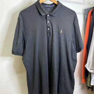 Mörkgrå pikétröja från Polo Ralph Lauren - Säljer en klassisk mörkgrå pikétröja från Polo Ralph Lauren med broderad logga på bröstet. Tröjan har krage, tre knappar och korta ärmar. Perfekt för en stilren och avslappnad look.