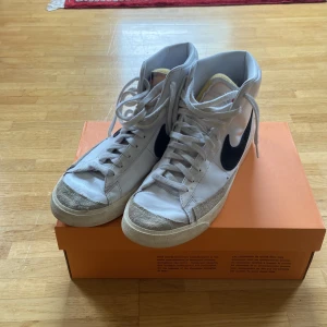 Nike Blazer Mid 77 Vintage vita sneakers - Säljer ett par klassiska Nike Blazer Mid 77 Vintage sneakers i vitt skinn med svarta swoosh-loggor på sidorna. Skorna har högt skaft, snörning och beige mockadetaljer vid tån och sidorna. Perfekta för dig som gillar retrostil!