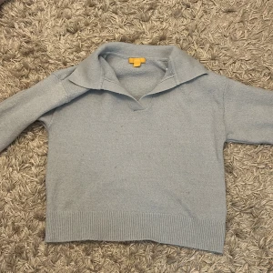 Ljusblå stickad tröja från Gina Tricot - Mysig ljusblå stickad tröja från Gina Tricot med krage och v-ringning. Tröjan har långa ärmar och bred ribbad kant nedtill och vid ärmsluten. Perfekt för kyligare dagar och enkel att matcha med jeans eller kjol.