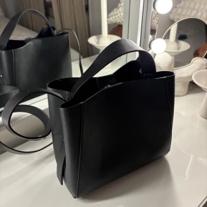 Svart axelväska i skinn - Stilren svart axelväska i slätt skinn med bred axelrem och handtag. Väskan har en minimalistisk design och passar perfekt för att bära dina viktigaste saker. Enkel att matcha med olika outfits. Från Gina. 