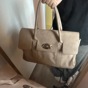 Beige handväska med gulddetalj - Säljer en stilren beige handväska med två handtag och guldfärgade vridknapp framtill. Väskan har klassisk form och är perfekt för dig som vill ha en rymlig men fin väska 
