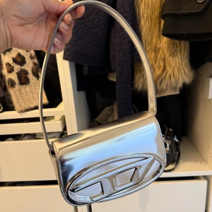 Diesel väska - Säljer en riktigt snygg axelväska i metallic silver från Diesel. Väskan har en unik oval form med ett stort dekorativt emblem på framsidan. Perfekt för att lyfta vilken outfit som helst med en futuristisk touch. Med väskan följer dustbag samt längre axelband med! 