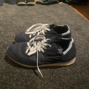 Phillips modell  - Säljer ett par mörkblå sneakers med vita snören och ljus sula. Skorna har en klassisk rund tå och platt sula, perfekt för en avslappnad stil. Materialet är syntet med textilkänsla och insidan är ljusgrå.