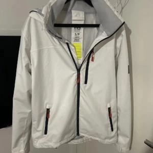 Vit vindjacka med huva - Snygg vit vindjacka från Helly Hansen med svart dragkedja och röda detaljer på fickorna. Jackan har huva med meshfoder och två sidofickor med dragkedja. Perfekt för blåsiga dagar.