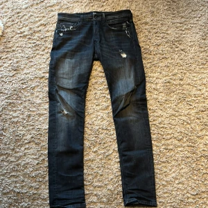 Svarta Replay jeans med slitningar - Säljer ett par svarta Replay jeans med snygga slitningar och detaljer framtill och baktill. Jeansen har fem fickor, knappgylf och slim ben. Iprincip nyskick knappt använda 