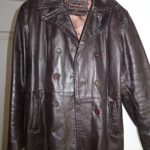 Brun skinnrock från Woodlands - Säljer en klassisk brun skinnrock från Woodlands med dubbelknäppning och bred krage. Jackan har långa ärmar och två fickor framtill. Perfekt för dig som gillar tidlös stil och vill ha något som sticker ut.