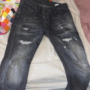 Svarta slitna jeans från Dsquared2 - Säljer ett par svarta jeans från Dsquared2 med slitningar och distressed detaljer framtill. Jeansen har klassisk femficksmodell, normal passform och knappgylf. Märkeslapp bak i vitt med röd logga.