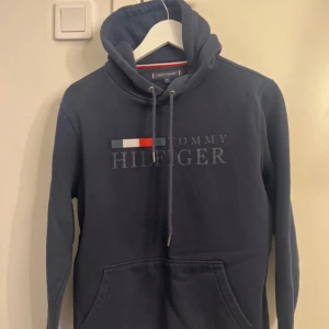 Mörkblå hoodie från Tommy Hilfiger - Säljer en mörkblå hoodie från Tommy Hilfiger med klassisk logga och broderad flagga på bröstet. Tröjan har huva med dragsko och en stor magficka. Perfekt för dig som gillar stilrena och bekväma plagg.
