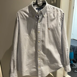 Randig skjorta från Ralph Lauren - Klassisk skjorta från Ralph Lauren med tunna blå och vita ränder, broderad logga på bröstet och knappar hela vägen. Skjortan har lång ärm och button-down krage, perfekt för en stilren look.