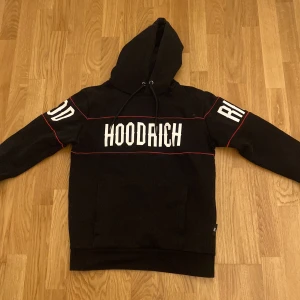 Svart hoodie från Hoodrich - Säljer en svart hoodie från Hoodrich med vit text på bröstet och ärmarna. Hoodien har en justerbar huva med dragsko och en stor ficka framtill. Den har röda detaljer längs med sömmarna som ger en snygg kontrast.