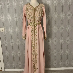 Rosa marockansk kaftan långklänning med guldbroderi - Elegant rosa marockansk kaftan långklänning med vackra guldbroderier och ett matchande bälte i midjan. Klänningen har långa ärmar och en v-ringad halslinje, perfekt för speciella tillfällen. Köpt från Spanien 