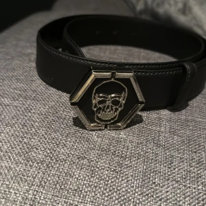 Svart bälte med dödskalle-spänne från Philipp Plein - Snyggt svart bälte i skinn från Philipp Plein med ett coolt dödskalle-spänne i silver. Perfekt för att ge din outfit en edgy touch. Tillverkat i Italien och kommer i originalförpackning. Längd 85cm