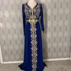 Blå marockansk kaftan långklänning med guldbroderi - Elegant blå kaftan långklänning med detaljerat guldbroderi längs framsidan och ärmarna. Klänningen har en v-ringad hals och ett matchande bälte i midjan som framhäver figuren. Perfekt för speciella tillfällen. 