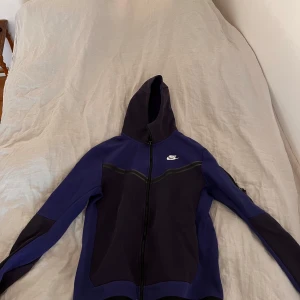 Nike tech fleece - Säljer en snygg blå och svart nike tech med dragkedja och huva. Perfekt för träning eller en avslappnad dag. Den har långa ärmar och en stilren design med Nike-logga på bröstet. Skriv till mig om du har några frågor eller funderingar 😃