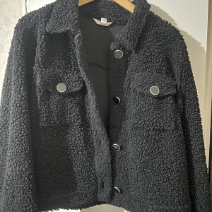 Svart teddyjacka med knappar - Mysig svart teddyjacka med stora knappar framtill. Jackan har två bröstfickor och en klassisk krage. Perfekt för kyliga dagar när du vill hålla dig varm och stilren.