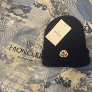 Svart stickad mössa från Moncler - Snygg svart stickad mössa från Moncler med deras ikoniska logga framtill. Perfekt för att hålla värmen under kyliga dagar. Enkel och stilren design som passar till det mesta. PRIS KAN DISKUTERAS 💯