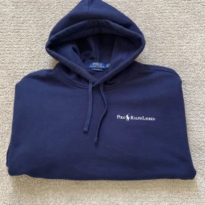 Mörkblå hoodie från Polo Ralph Lauren - Säljer en snygg mörkblå hoodie från Polo Ralph Lauren med broderad logga på bröstet. Hoodien har en avslappnad passform och en praktisk känguruficka framtill. Perfekt för en stilren och bekväm look.