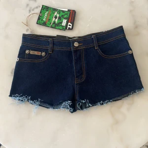 Mörkblå lågmidjade jeansshorts från Diesel - Snygga mörkblå lågmidjade jeansshorts från Diesel med fransiga kanter och coola broderade detaljer på bakfickorna. Perfekta för en avslappnad stil. De har en klassisk femficksdesign och knappgylf. Dem passar inte så inga bilder med dem på❤️ midjemått 37-38 cm rakt över