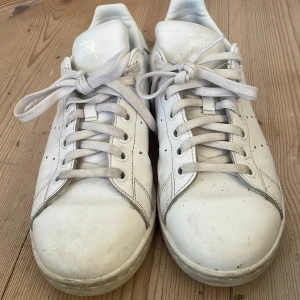 Vita Adidas Stan Smith sneakers - Säljer ett par klassiska vita Adidas Stan Smith sneakers. Skorna har en stilren design med snörning och perforerade detaljer på sidorna. Perfekta för en avslappnad och trendig look.Dock har den lite slitning på sidorna som ni kan se på bilden och har lite stänk av något.Tycker inte att det påverkar din still men vet inte om det kan gå bort.