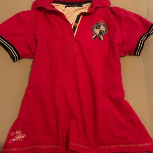 Rosa piké tshirt  - Snygg rosa piké tshirt med broderad logga på bröstet och textdetalj vid fållen. T-shirten har en klassisk pikékrage och knappar framtill. Perfekt för en avslappnad och stilren look.