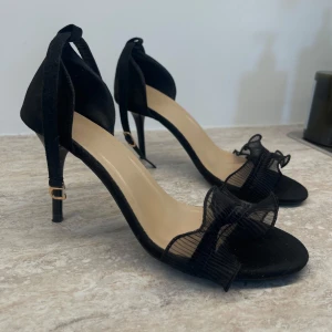 Svarta klackskor - Eleganta svarta klackskor med öppen tå och volanger framtill. De har ett  justerbart band runt ankeln.