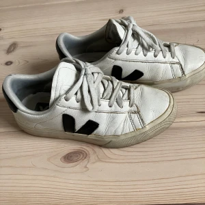 Vita sneakers från Veja storlek 36 - Säljer ett par vita sneakers från Veja med svarta detaljer. Skorna har snörning och en klassisk rund tå. Perfekta för en stilren look. Innermått ca 22,5.