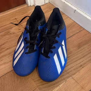 Blå fotbollsskor från Adidas - Snygga blå fotbollsskor från Adidas med vita ränder och svarta detaljer. Skorna har snörning och en slitstark sula med dobbar för bra grepp på planen. Perfekta för fotbollsträning och matcher.