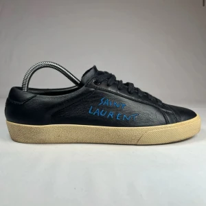 Svarta sneakers från Saint Laurent - Snygga svarta sneakers från Saint Laurent med blå broderad logga på sidan. Skorna har en klassisk design med rund tå och svart snörning. Yttersulan är i en kontrasterande beige färg som ger en stilren look. Ny pris 8299kr