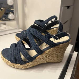 Jeansfärgade kilklackssandaler  - Snygga jeansfärgade sandaler med kilklack och flätad sula. Storlek 38.  De har flera remmar som korsar foten och en justerbar rem runt ankeln. Perfekta för en somrig look! Använda fåtal gånger.