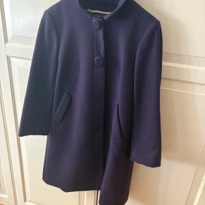 Mörklila kappa med knappar - Finns på Kungsholmen. Elegant mörklila kappa med knappar framtill och diskreta fickor. 