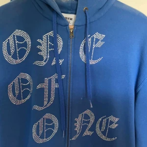 Blå hoodie från One of One - Snygg blå hoodie från One of One med dragkedja och text i glittriga stenar på framsidan. Perfekt för en cool och avslappnad stil. Den har en klassisk känguruficka och justerbar huva med dragsko.