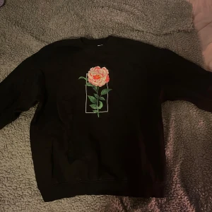 Svart sweatshirt med blomtryck och turnélista - Säljer en svart sweatshirt med en stor blomma tryckt på framsidan och en turnélista på baksidan. Perfekt för fans av musik och stil. Tröjan har långa ärmar och en bekväm passform.