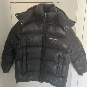 Svart pufferjacka från Axel Arigato - Säljer en svart pufferjacka från Axel Arigato med dragkedja och hög krage. Jackan har en luva och är långärmad, perfekt för kyliga dagar. Tillverkad i återvunnen polyester för en hållbar stil. Märket 'Arigato 2014' är tryckt på bröstet. Små hål i fickorna som kan lagas vid behov.