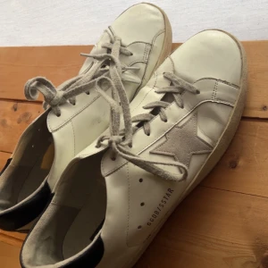 Vita sneakers från Golden Goose - Snygga vita sneakers från Golden Goose med en ikonisk stjärndetalj på sidan. Skorna har en klassisk snörning och en kontrasterande svart häl. Perfekta för en stilren look. Storlek 43 men passar även 42. Skick 7/10. Säljs då de inte kommer till användning. Priset är inte hugget i sten! Hör av er om ni har frågor😊⭐️