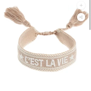 Beige armband med text - Beige armband med texten ”C'est La Vie” broderad i vitt. Armbandet har tofsar i ändarna och är justerbart för att passa olika storlekar. Helt oanvänd med ett ordinarie pris på 285kr. 