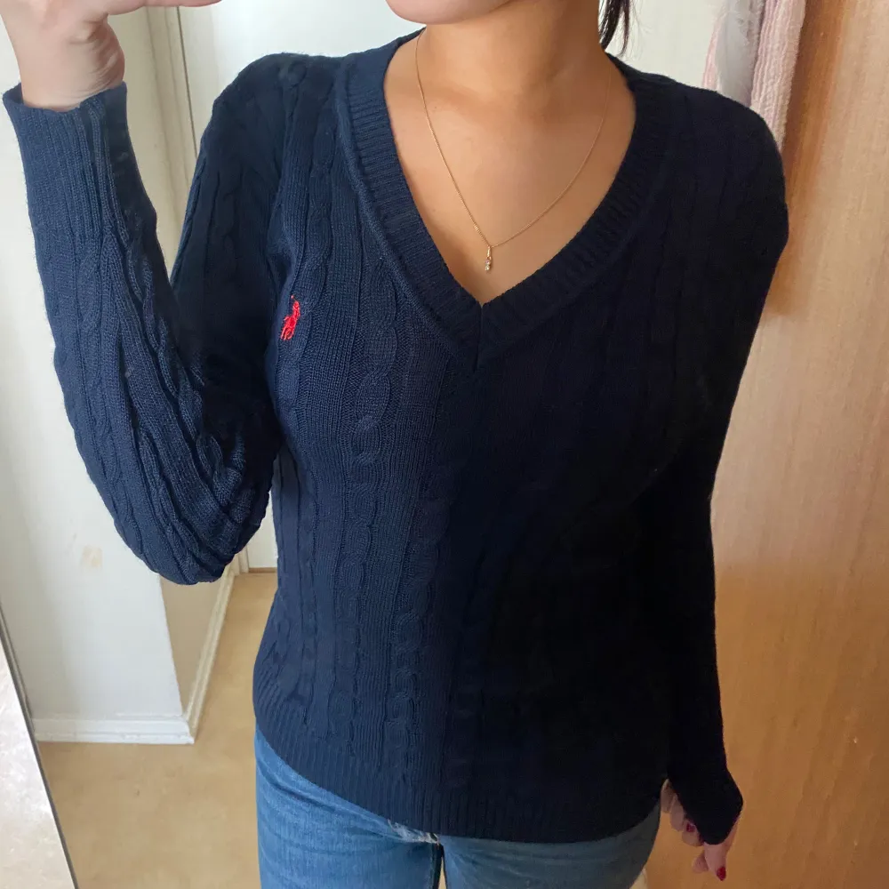 Säljer en stilren mörkblå stickad tröja från Ralph Lauren med v-ringning och ett diskret rött logomärke på bröstet. Tröjan har långa ärmar och en klassisk kabelstickad design. Perfekt för en avslappnad men ändå elegant look.. Neuletakit & Villapaidat.