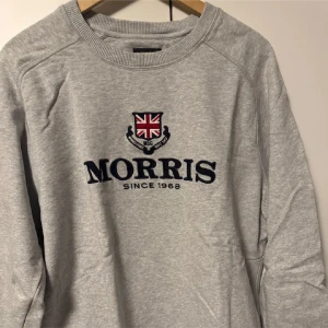 Grå och marinblå tröja från Morris - Två stilrena tröjor från Morris med broderad text och brittisk flagga. Den grå tröjan har texten 'Morris Since 1968' medan den marinblå har 'Morris The Gent Est TM Since 1968 England'. Perfekta för en avslappnad och klassisk look.