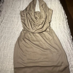 Beige klänning från ICHI - Snygg beige klänning från ICHI med draperad halslinje. Perfekt för en stilren look. Klänningen har en elegant passform och är tillverkad i ett mjukt skimrande material.