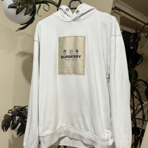 Vit hoodie från Burberry - Säljer en stilren vit hoodie från Burberry🥳 med en beige patch på bröstet. Hoodien har långa ärmar och en klassisk huva med dragsko. Perfekt för en avslappnad och trendig look. Det ni ser på bilden är det ni får😁 ett plagg som är köpt i bulk så jag vet lila lite som ni, men jag har kollat så gott jag kan på alla möjliga sajter och det jag kan kolla stämmer överens med originalet väldigt bra tröja raka o fina sömmar, kvalitets lappar och tyg