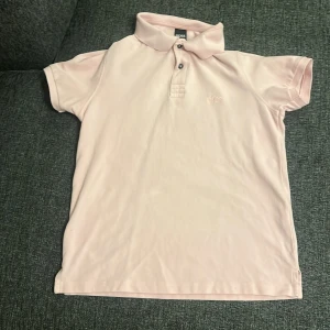 Ljusrosa pikétröja från Hugo Boss - Säljer en stilren ljusrosa pikétröja från Hugo Boss. Tröjan har en klassisk krage med knappar och är kortärmad. Perfekt för en avslappnad men ändå snygg look. Har används 2 gånger max. Storlek: (Small)