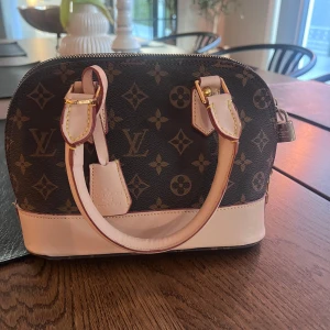 Brun handväska  - Säljer en elegant handväska. Brunt och beige med det klassiska monogrammönstret. Väskan har två handtag och en dragkedja upptill. Finns även en innerficka. Perfekt för att ge en lyxig touch till din outfit.