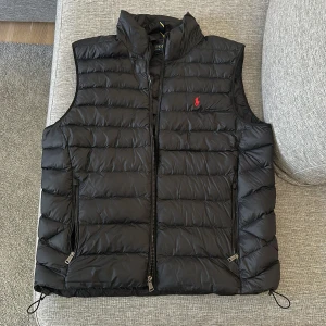 Svart dunväst från Ralph Lauren - Säljer en stilren svart dunväst från Ralph Lauren med dragkedja och broderad logga på bröstet. Perfekt för lager-på-lager under kyliga dagar. Västen har en hög krage och justerbara dragskor i nederkant för bästa passform.